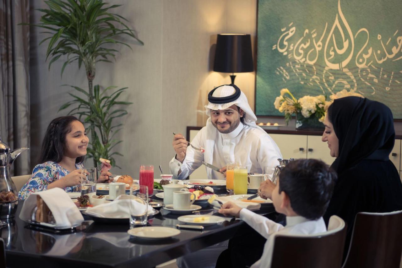 מלון מכה Jabal Omar Hyatt Regency Makkah מראה חיצוני תמונה The photo depicts a cheerful family gathered around a dining table for a meal. They appear to be enjoying breakfast or a brunch. The table is set with various dishes and drinks, including juices and plates of food. 

A man wearing traditional attire 