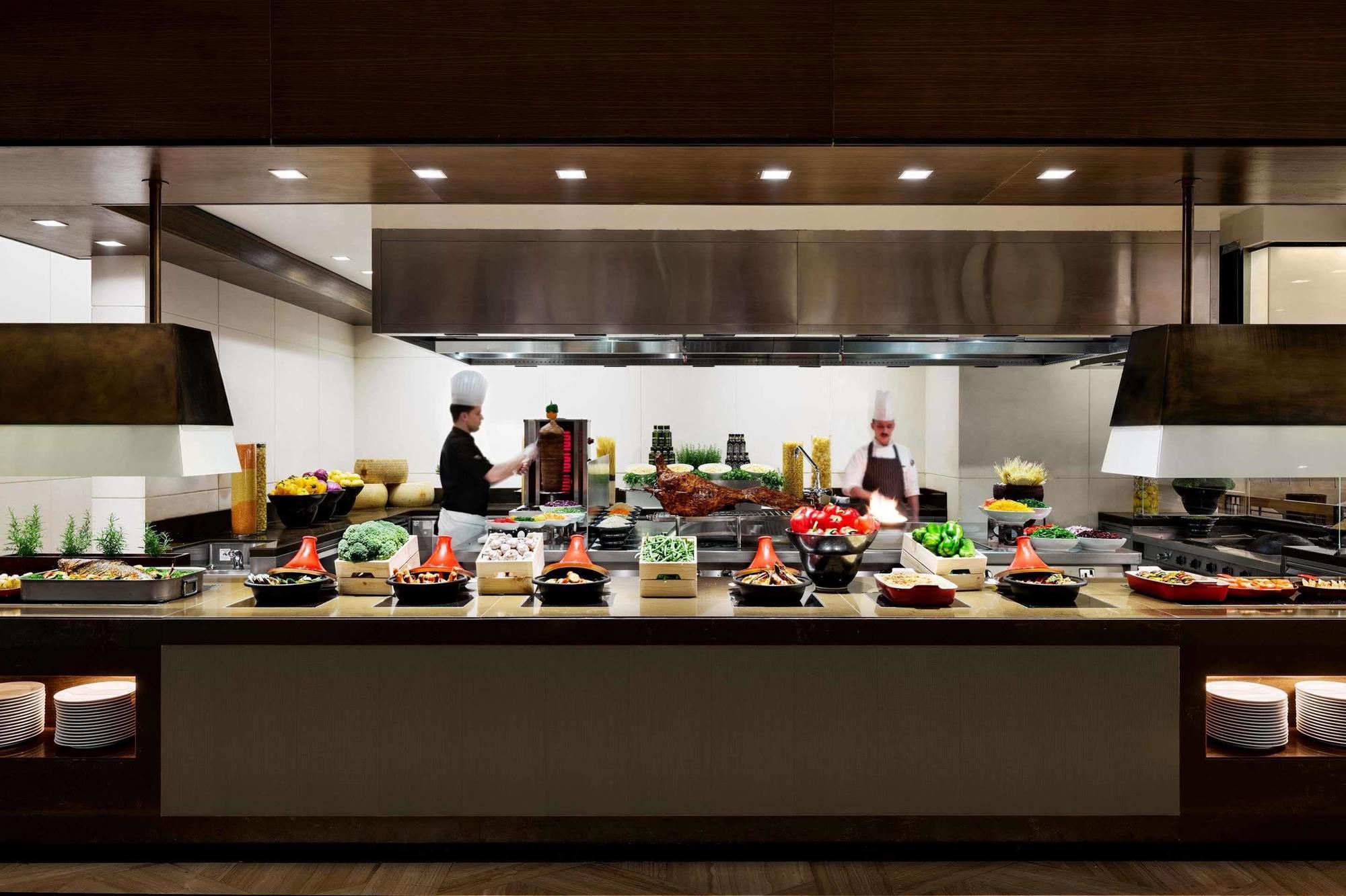 מלון מכה Jabal Omar Hyatt Regency Makkah מראה חיצוני תמונה The photo shows a well-organized kitchen or buffet area. In the foreground, there are several food stations displaying a variety of dishes, including fresh vegetables, meats, and other culinary items arranged in bowls or trays. Behind the counter, tw
