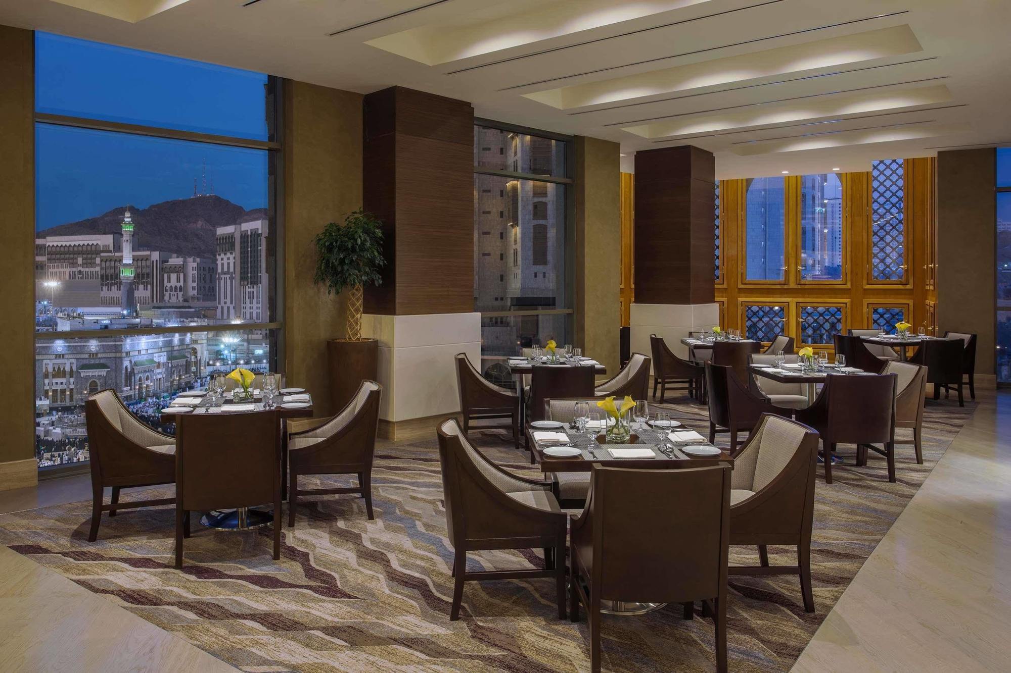 מלון מכה Jabal Omar Hyatt Regency Makkah מראה חיצוני תמונה The photo shows a sophisticated restaurant interior, featuring elegant round tables with high-backed chairs arranged neatly. The space is well-lit with large windows that provide a view of the city skyline at dusk, with mountains in the background. T
