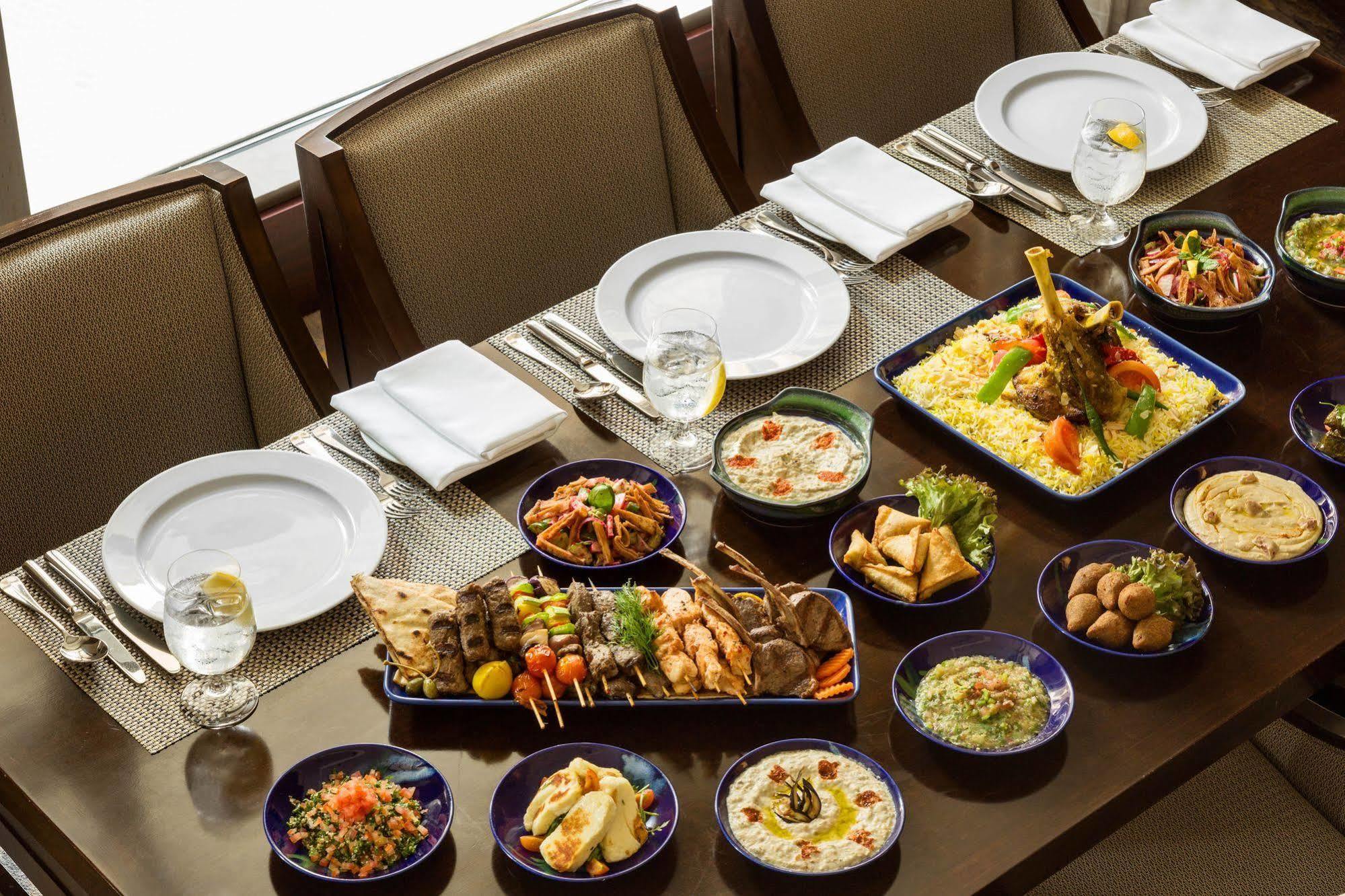 מלון מכה Jabal Omar Hyatt Regency Makkah מראה חיצוני תמונה The photo shows a beautifully arranged dining table set for a feast. There are several plates and dishes filled with a variety of foods:

1. **Main Dishes**: A large platter features grilled meats, including skewers and seasoned chicken, garnished wi