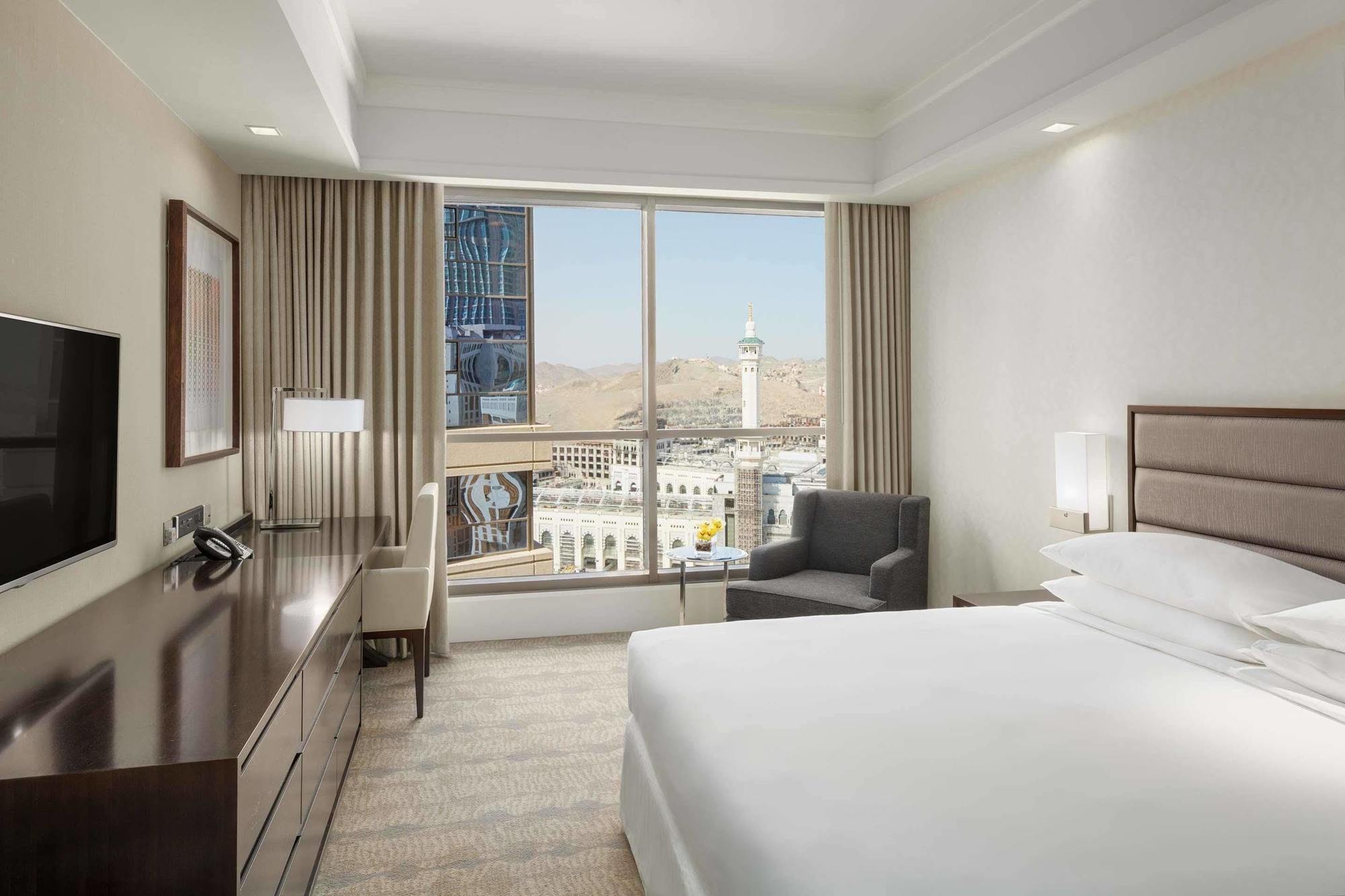 מלון מכה Jabal Omar Hyatt Regency Makkah מראה חיצוני תמונה The photo shows a modern hotel room featuring a large window that provides a view of an outdoor landscape, likely part of a city or landmark. The room includes a bed with white linens, a side table with a lamp, and a small seating area with a chair a