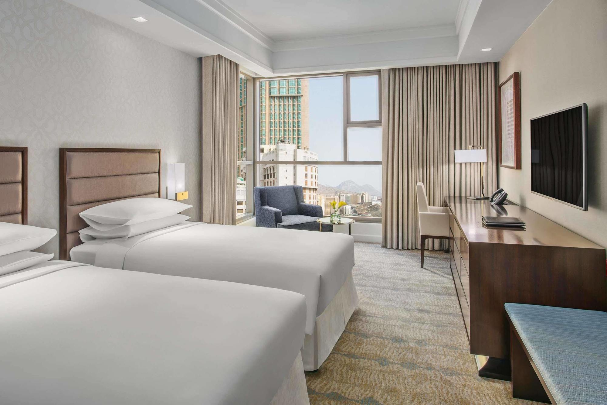 מלון מכה Jabal Omar Hyatt Regency Makkah מראה חיצוני תמונה The photo shows a modern hotel room featuring two single beds with white bedding. There's a spacious window allowing natural light to fill the room, which is adorned with light-colored curtains. In one corner, there's a comfortable armchair next to a